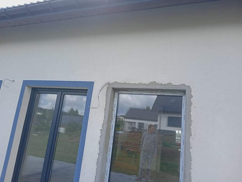 Ściana zewnętrzna budynku z dwoma oknami, jedno z niebieską ramą i drugie w trakcie uszczelniania, z widocznym odbiciem osoby w szybie.