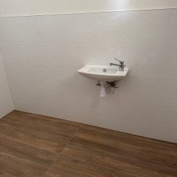 Ergo Grzegorz Sztyma - Biała umywalka zamontowana na ścianie z dekoracyjnymi, trójwymiarowymi płytkami. Podłoga wyłożona brązowymi panelami imitującymi drewno.