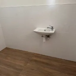 Biała umywalka zamontowana na ścianie z dekoracyjnymi, trójwymiarowymi płytkami. Podłoga wyłożona brązowymi panelami imitującymi drewno.