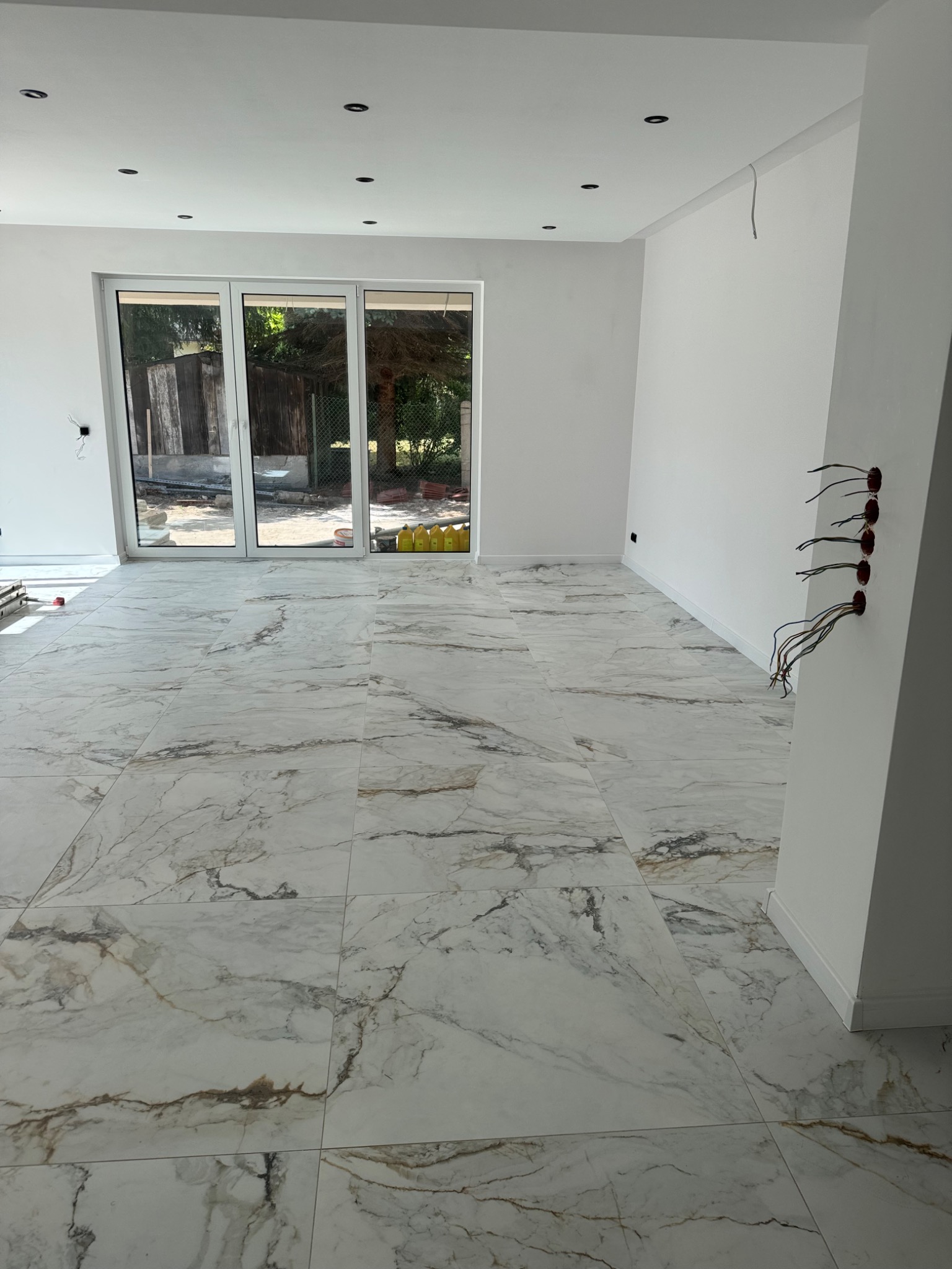 Wykończone podłogi z marmurowych płytek w jasnym wnętrzu, widoczne puszki elektryczne i przygotowane kable. W tle okno z widokiem na zieleń.