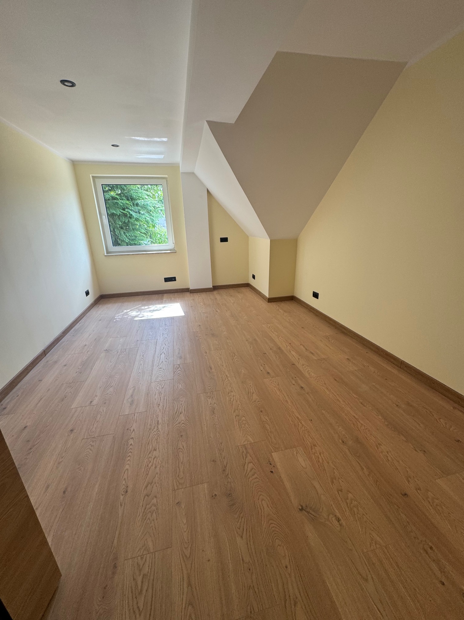 Jasne wnętrze poddasza z drewnianą podłogą i oknem. Ściany w odcieniach beżu, widoczne gniazdka elektryczne. Ukośny sufit dodaje przestrzeni unikalnego charakteru.