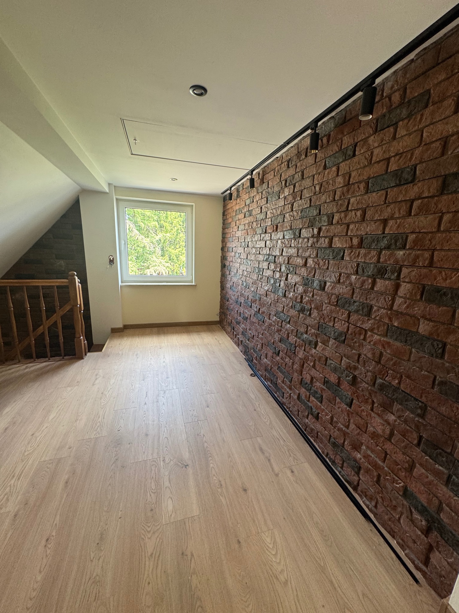 Poddasze po remoncie: drewniana podłoga, ściana z cegły z oświetleniem szynowym. Widoczne okno i fragment schodów. Całość w jasnych, naturalnych barwach.