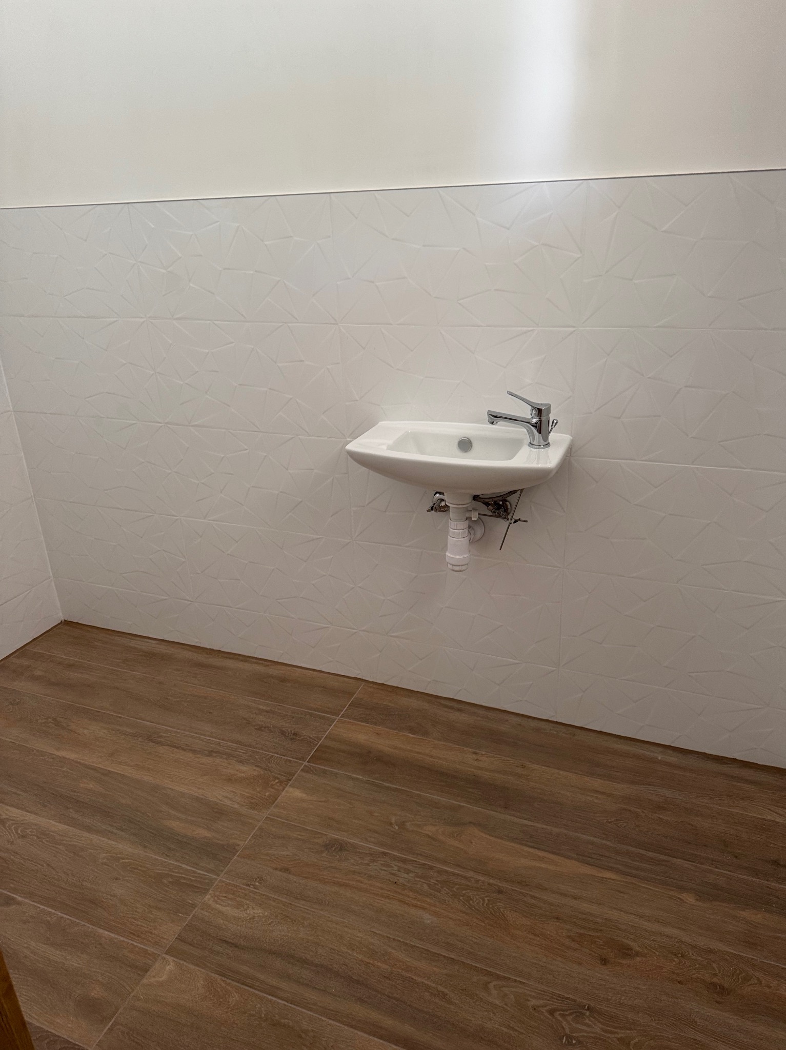 Biała umywalka zamontowana na ścianie z dekoracyjnymi, trójwymiarowymi płytkami. Podłoga wyłożona brązowymi panelami imitującymi drewno.