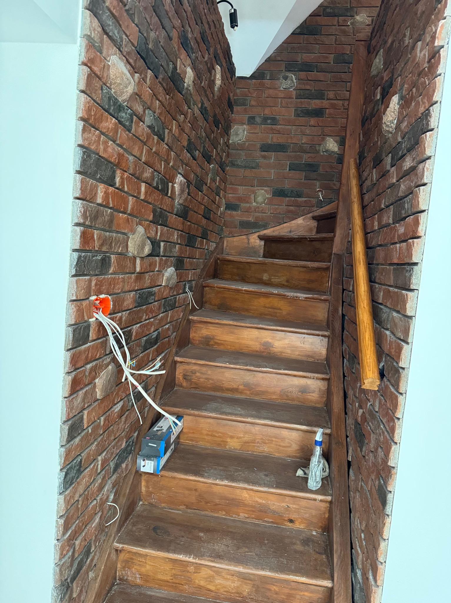 Klatka schodowa z drewnianymi schodami i ceglaną ścianą. Widoczne przewody elektryczne wystające ze ściany. Na schodach leżą pudełka i butelka ze spryskiwaczem.