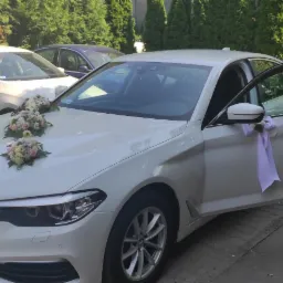 Białe BMW serii 5 udekorowane kwiatami i wstążkami, z otwartymi drzwiami, zaparkowane na chodniku przed budynkiem, przygotowane do ślubu.