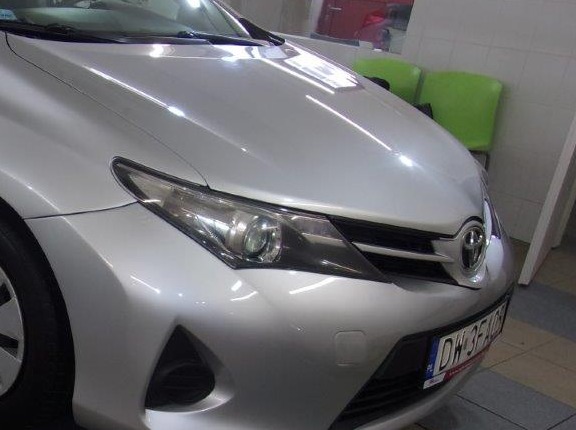 Przedni fragment srebrnego samochodu osobowego, z widocznym reflektorem, grillem z logo i fragmentem tablicy rejestracyjnej, sfotografowany w pomieszczeniu z zielonymi krzesłami w tle.