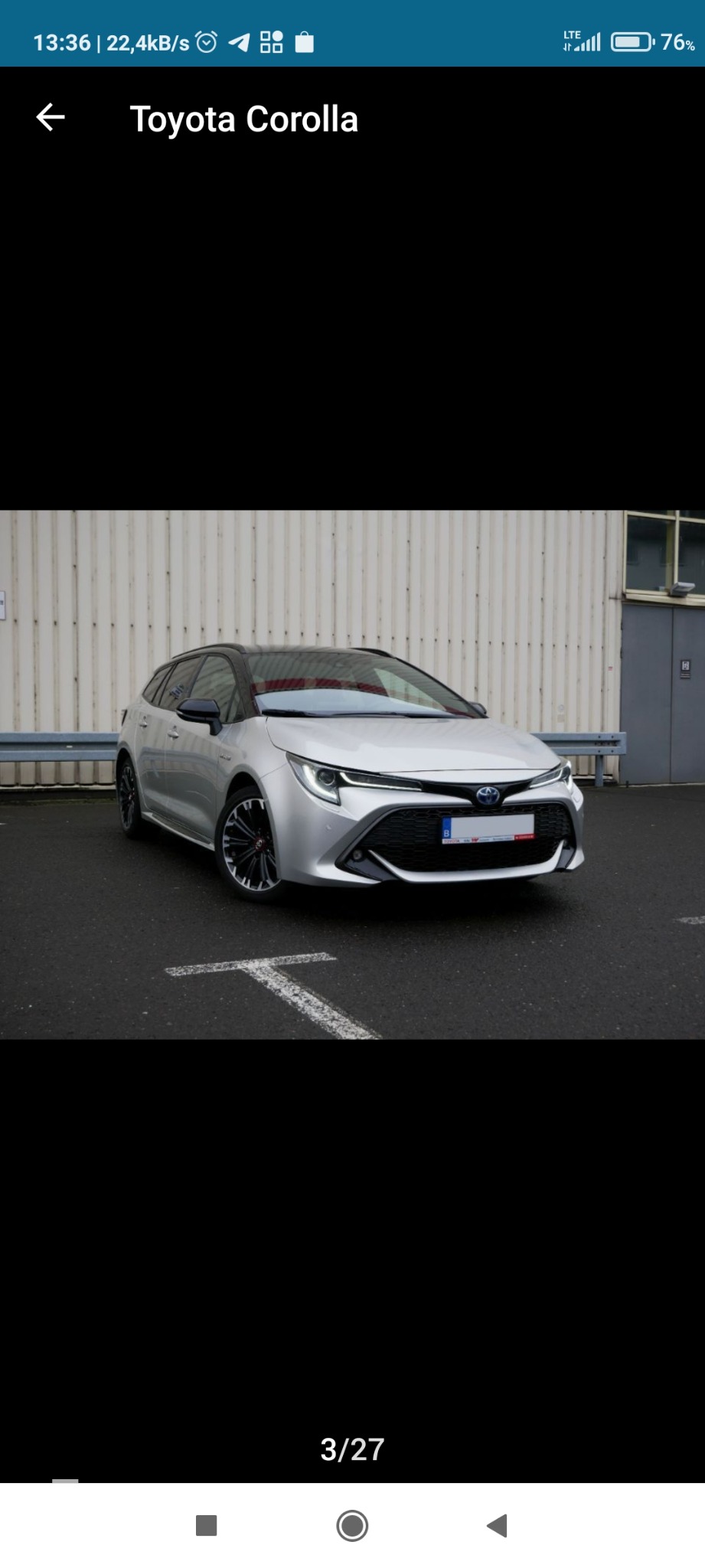 Srebrna Toyota Corolla kombi, zaparkowana na asfalcie z białymi oznaczeniami, na tle białej, blaszanej ściany budynku.