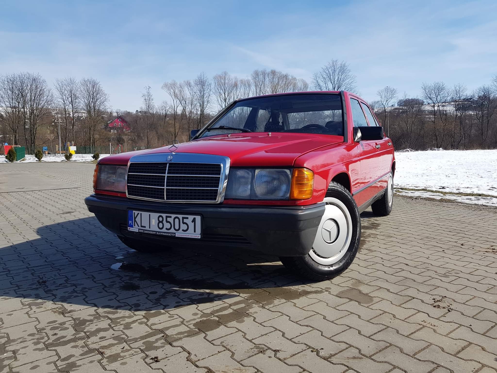 Czerwony Mercedes-Benz 190E na tle szarej kostki brukowej, z fragmentami śniegu w tle i błękitnym niebem.