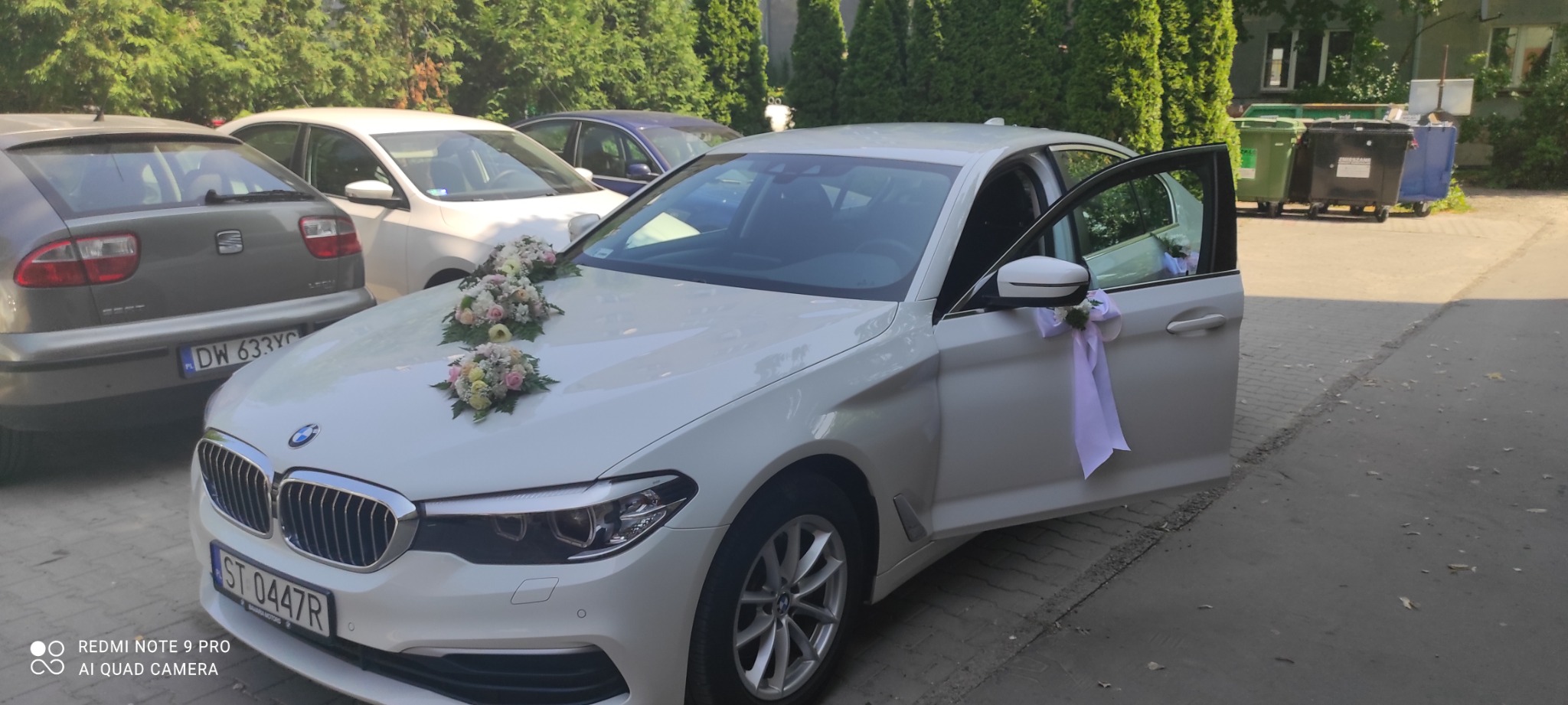 Białe BMW serii 5 udekorowane kwiatami i wstążkami, z otwartymi drzwiami, zaparkowane na chodniku przed budynkiem, przygotowane do ślubu.