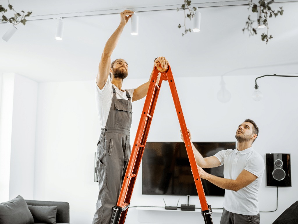 Dwóch elektryków montuje oświetlenie szynowe w salonie w Dąbrówce, jeden na drabinie w kombinezonie, drugi obserwuje z dołu.