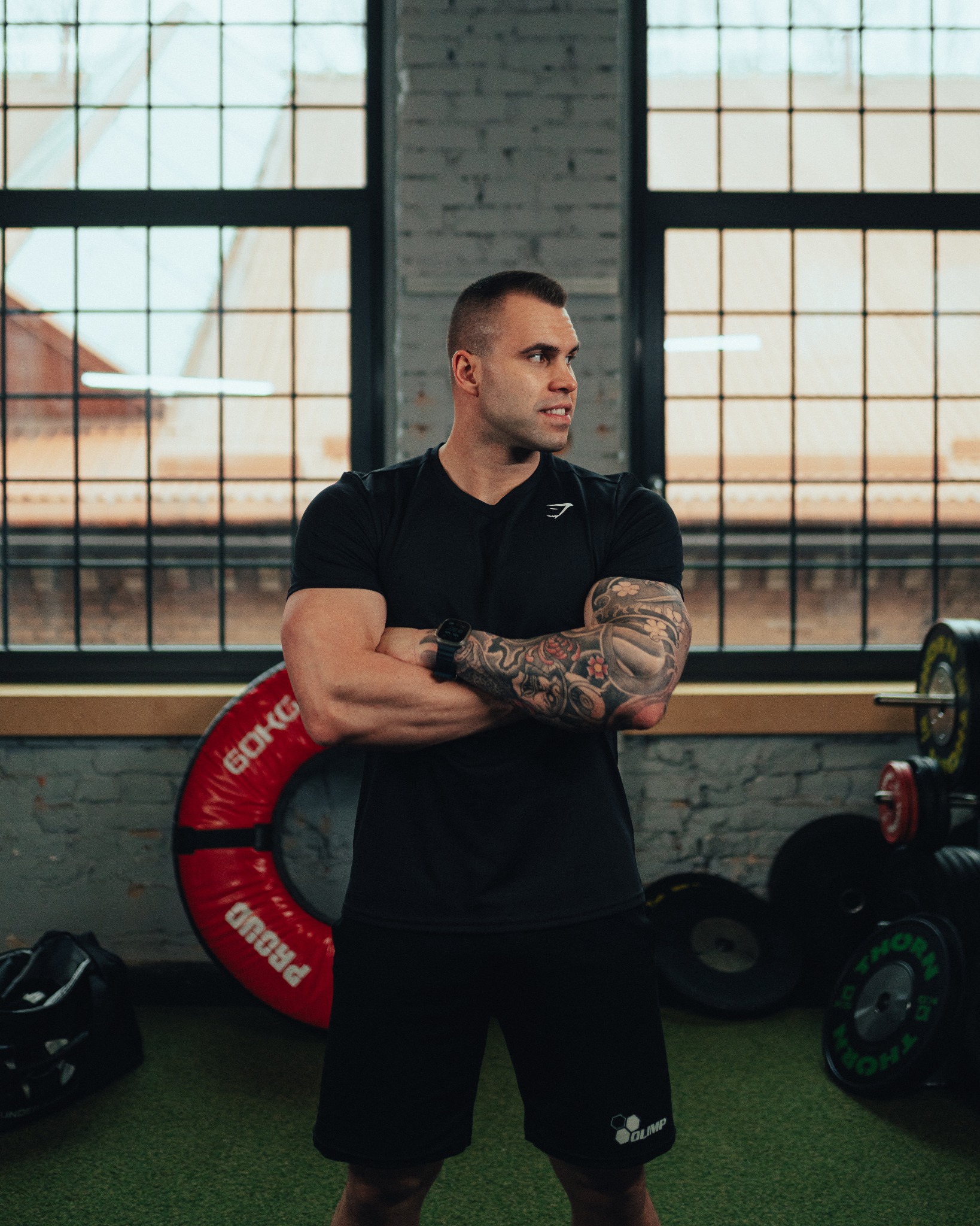 Mężczyzna z tatuażami na ramionach, ubrany w czarny strój sportowy, stoi z założonymi rękami na tle siłowni z dużym oknem i sprzętem do ćwiczeń.