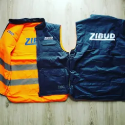 Dwie kamizelki robocze: jedna otwarta, z pomarańczowym wnętrzem i szarymi pasami odblaskowymi, druga granatowa z białym napisem 'ZIBUD' na plecach, leżące na drewnianej podłodze.