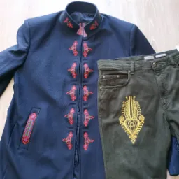 Granatowa zapinana kurtka z ludowym haftem kwiatowym zestawiona z oliwkowymi spodniami jeansowymi z haftowanym motywem na nogawce, leżące na drewnianej podłodze.