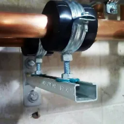 Miedziana rura przymocowana do ściany za pomocą metalowej obejmy z czarną gumową osłoną i srebrnego profilu montażowego typu C, zabezpieczonego nakrętkami i podkładkami w kolorze turkusowym.