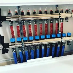 Rozdzielacz ogrzewania podłogowego z rurami w kolorze czerwonym i niebieskim, zamontowany w białej szafce, widoczne zawory i manometr.