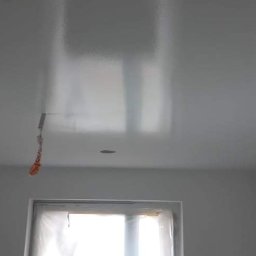 MaraBud - Biały, błyszczący sufit po malowaniu, widoczne odbicia światła, okno zabezpieczone folią malarską, w rogu zwisa sznurek z taśmy malarskiej, widoczny otwór pod oświetlenie.