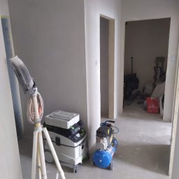 MaraBud - Wnętrze w trakcie prac wykończeniowych: ściany po gładziowaniu, widoczny kompresor, odkurzacz przemysłowy Festool oraz lampa na statywie.