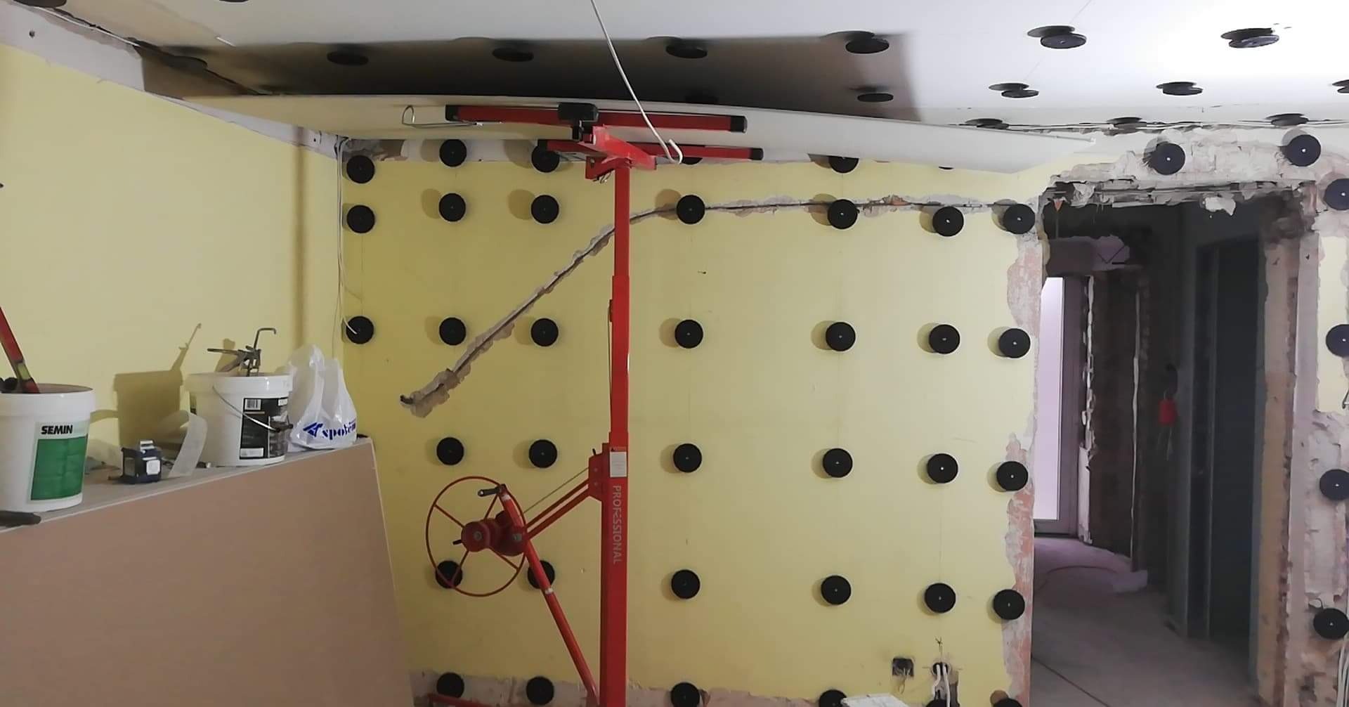 Montaż płyt karton gips w Białymstoku: żółta ściana z czarnymi mocowaniami, widoczny podnośnik do płyt i częściowo zamontowany sufit podwieszany.
