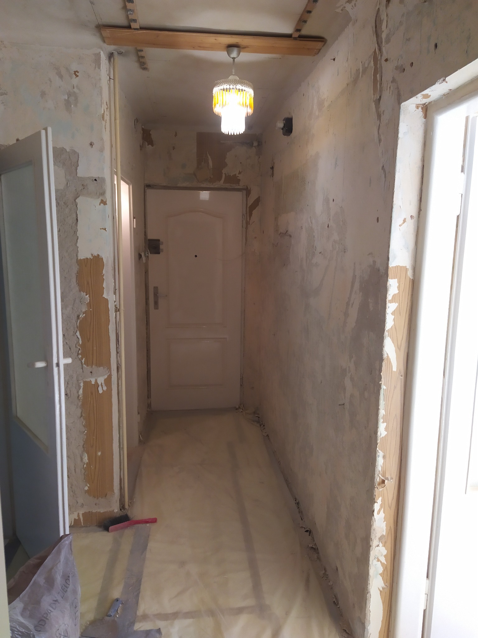 Korytarz w mieszkaniu w trakcie przygotowania do remontu, ściany częściowo obdarte z tapet, podłoga zabezpieczona folią, widoczny fragment drewnianej belki na suficie i wisząca lampa.
