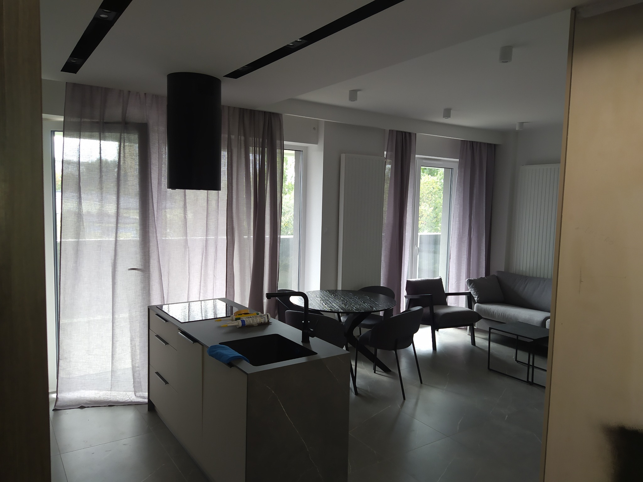 Nowoczesne wnętrze mieszkania z aneksem kuchennym, szarymi zasłonami i czarnymi akcentami oświetleniowymi i meblowymi, widoczne ślady prac wykończeniowych na blacie kuchennym.