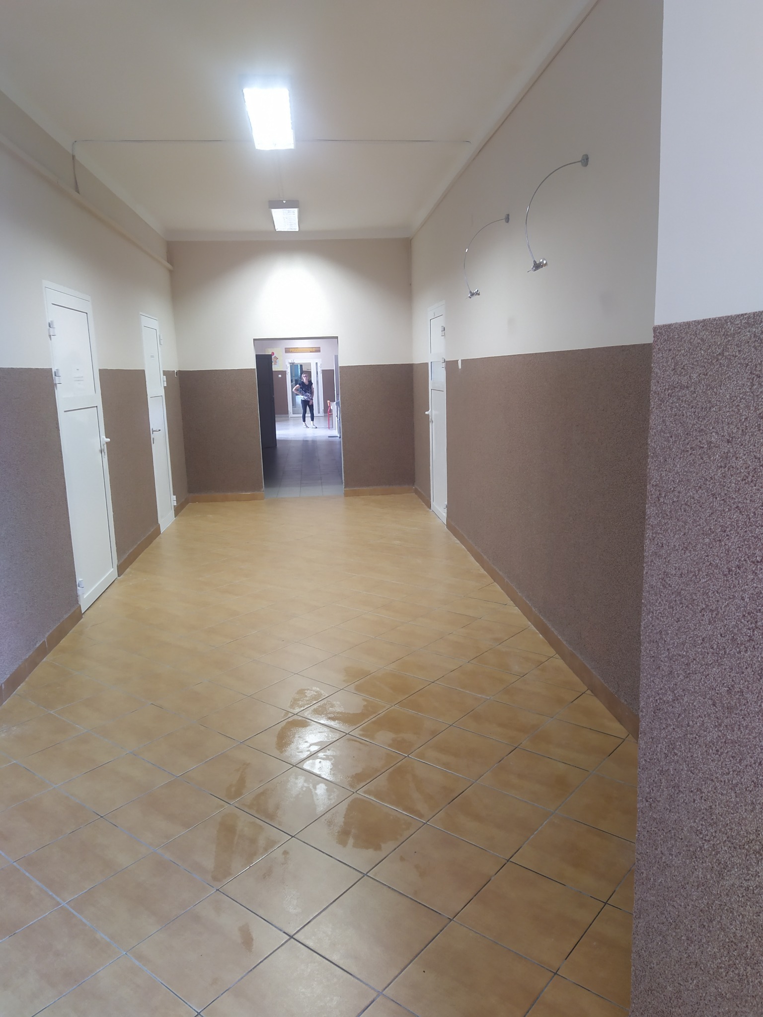 Długi korytarz z beżowymi płytkami podłogowymi, ściany podzielone na dwie części: górna pomalowana na jasny beż, dolna wykończona brązowym tynkiem strukturalnym, białe drzwi po obu stronach...