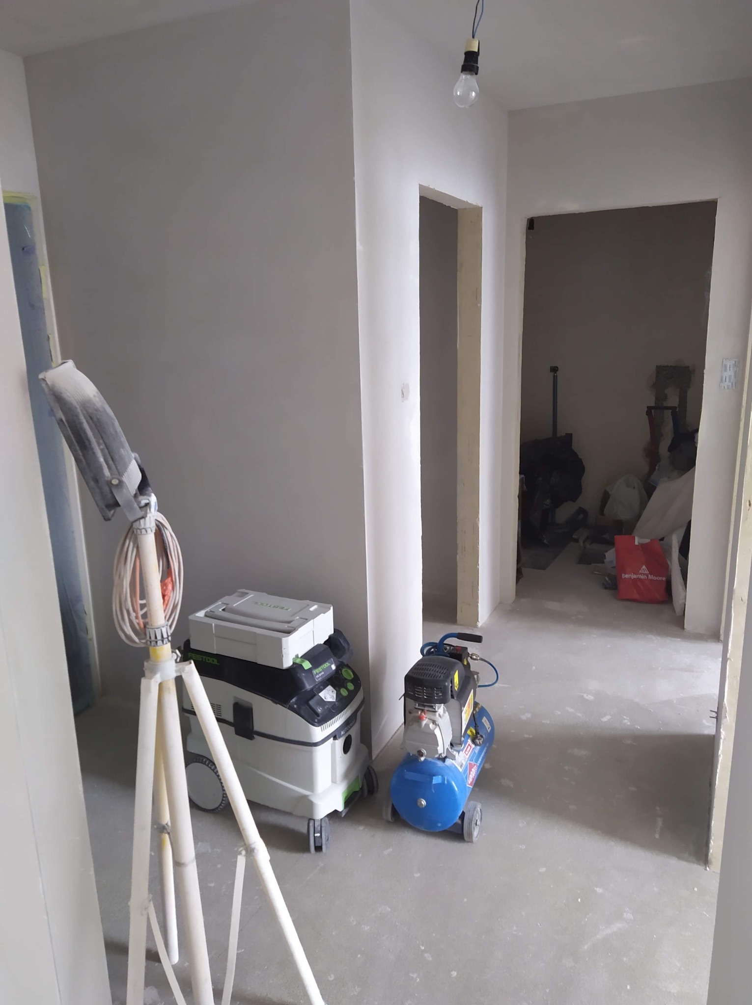 Wnętrze w trakcie prac wykończeniowych: ściany po gładziowaniu, widoczny kompresor, odkurzacz przemysłowy Festool oraz lampa na statywie.