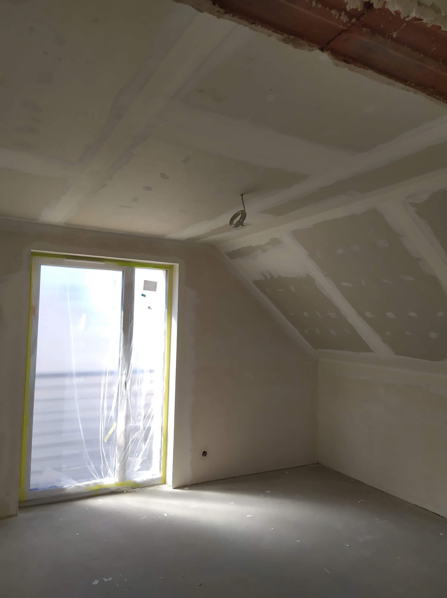 Poddasze w trakcie prac wykończeniowych: zamontowane płyty kartonowo-gipsowe na skośnym suficie i ścianach, okno zabezpieczone folią malarską, widoczne przewody elektryczne.