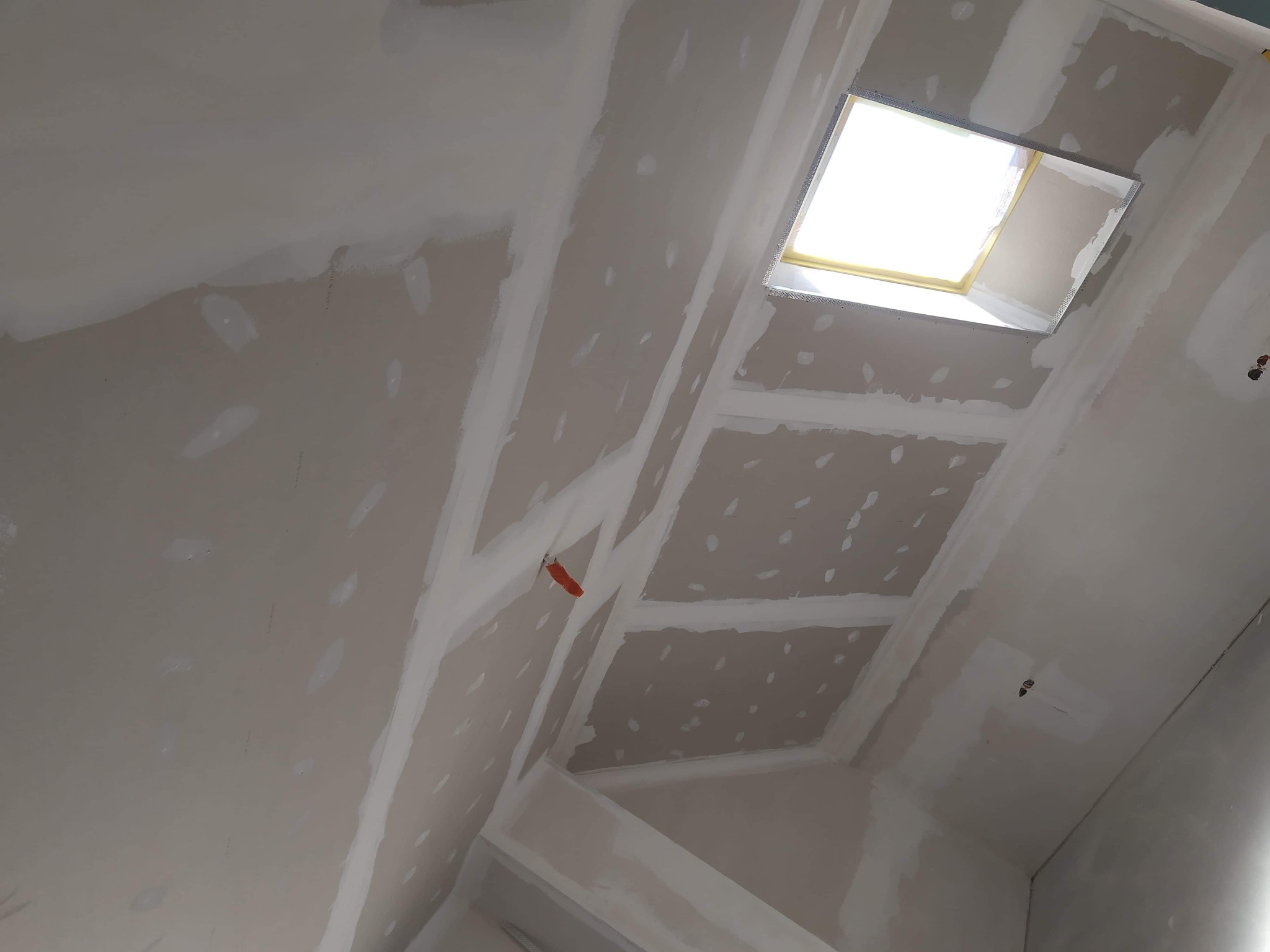 Skośny sufit poddasza z oknem dachowym w trakcie szpachlowania łączeń płyt kartonowo-gipsowych, widoczne ślady po wkrętach i pomarańczowy kabel.