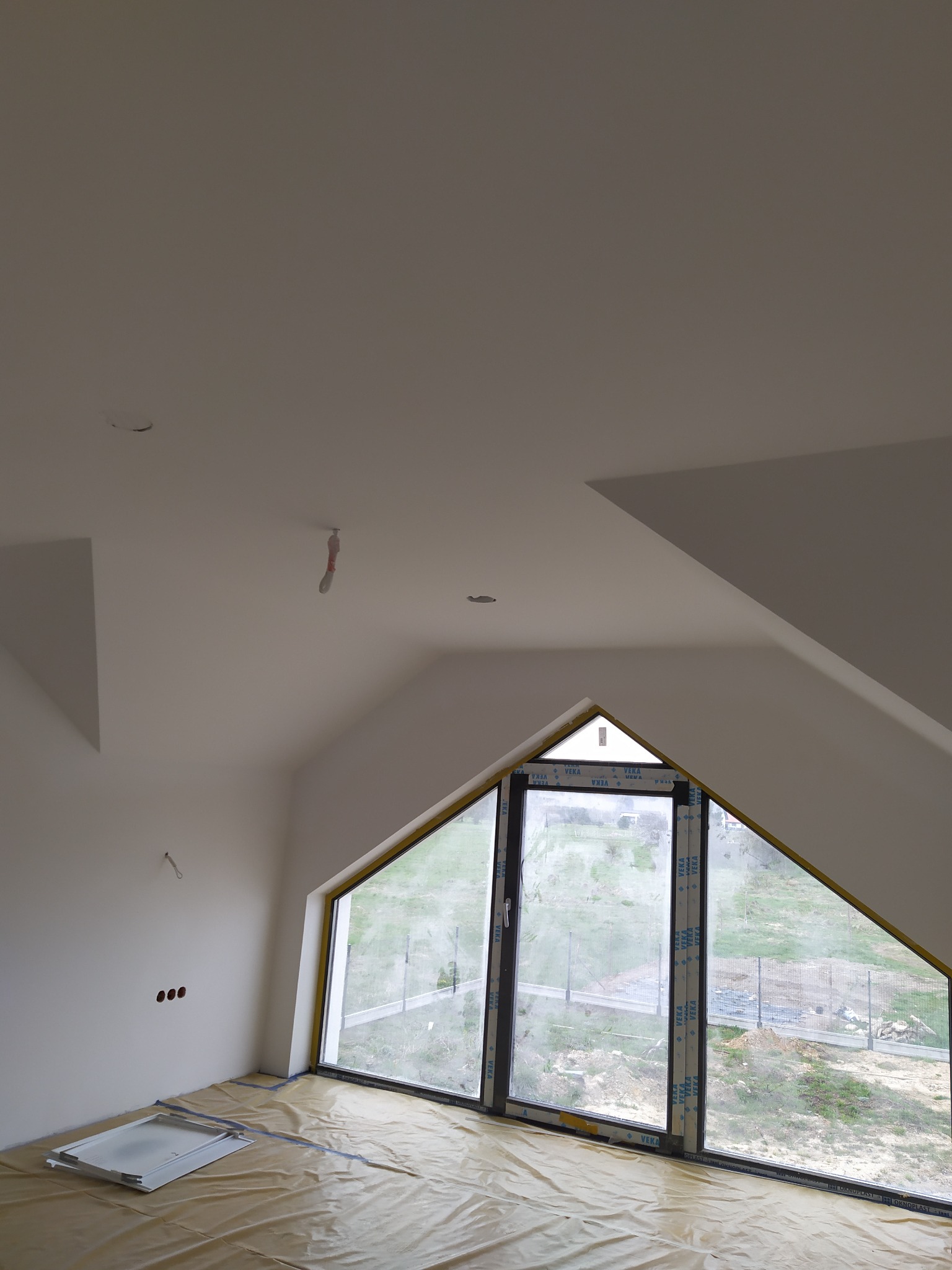 Poddasze ze skośnym oknem w kształcie trójkąta, folia ochronna na podłodze, przygotowane do prac wykończeniowych, widoczne puszki elektryczne w ścianie.
