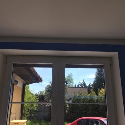 Sebud - Świeżo zamontowane, białe okno PCV z widocznymi śladami taśmy malarskiej zabezpieczającej, na tle granatowej ściany i widoku na ogród z czerwonym samochodem w odbiciu szyby.