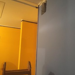 Sebud - Ściana w trakcie malowania na szaro, z widocznym fragmentem żółtej ściany, zabezpieczonej taśmą malarską, oraz drabina malarska w pomieszczeniu.