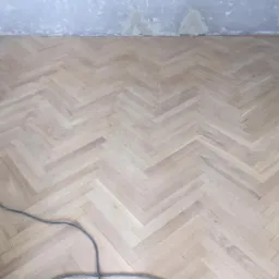 Ułożona parkietowa podłoga w jodełkę, jasne drewno, ściana w tle częściowo wyszpachlowana, widoczny kabel urządzenia do obróbki drewna.