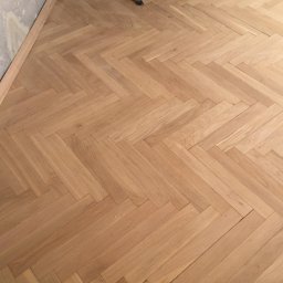 Sebud - Ukończona podłoga z parkietu ułożonego w jodełkę, jasne drewno, widoczna listwa przypodłogowa i fragment surowej ściany w rogu.