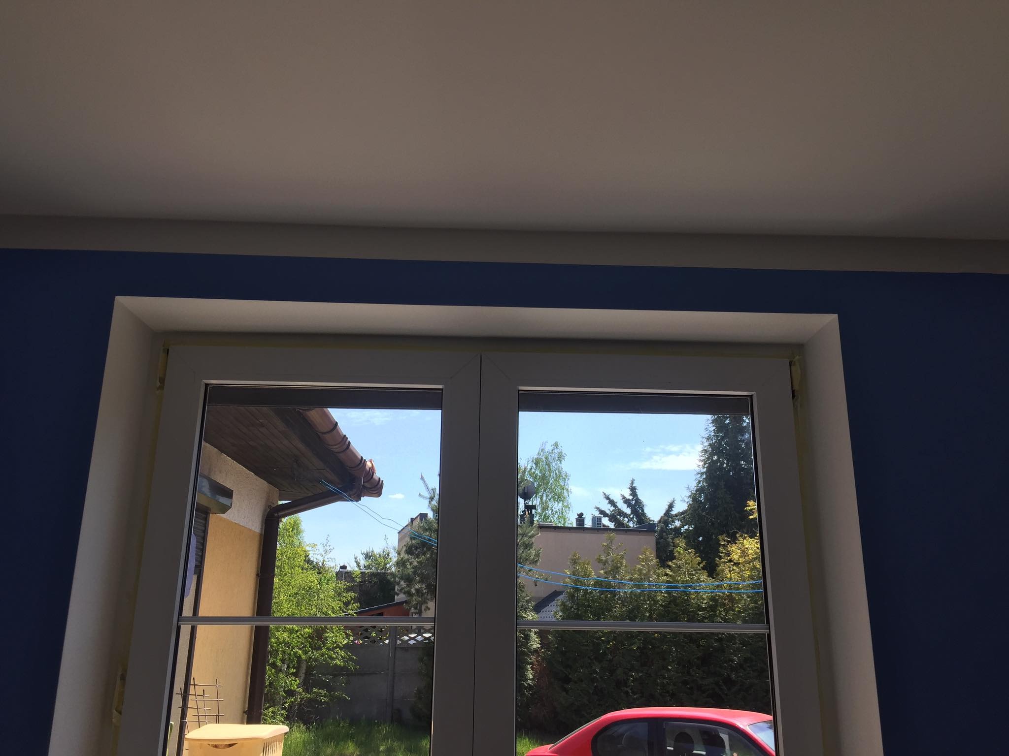 Świeżo zamontowane, białe okno PCV z widocznymi śladami taśmy malarskiej zabezpieczającej, na tle granatowej ściany i widoku na ogród z czerwonym samochodem w odbiciu szyby.