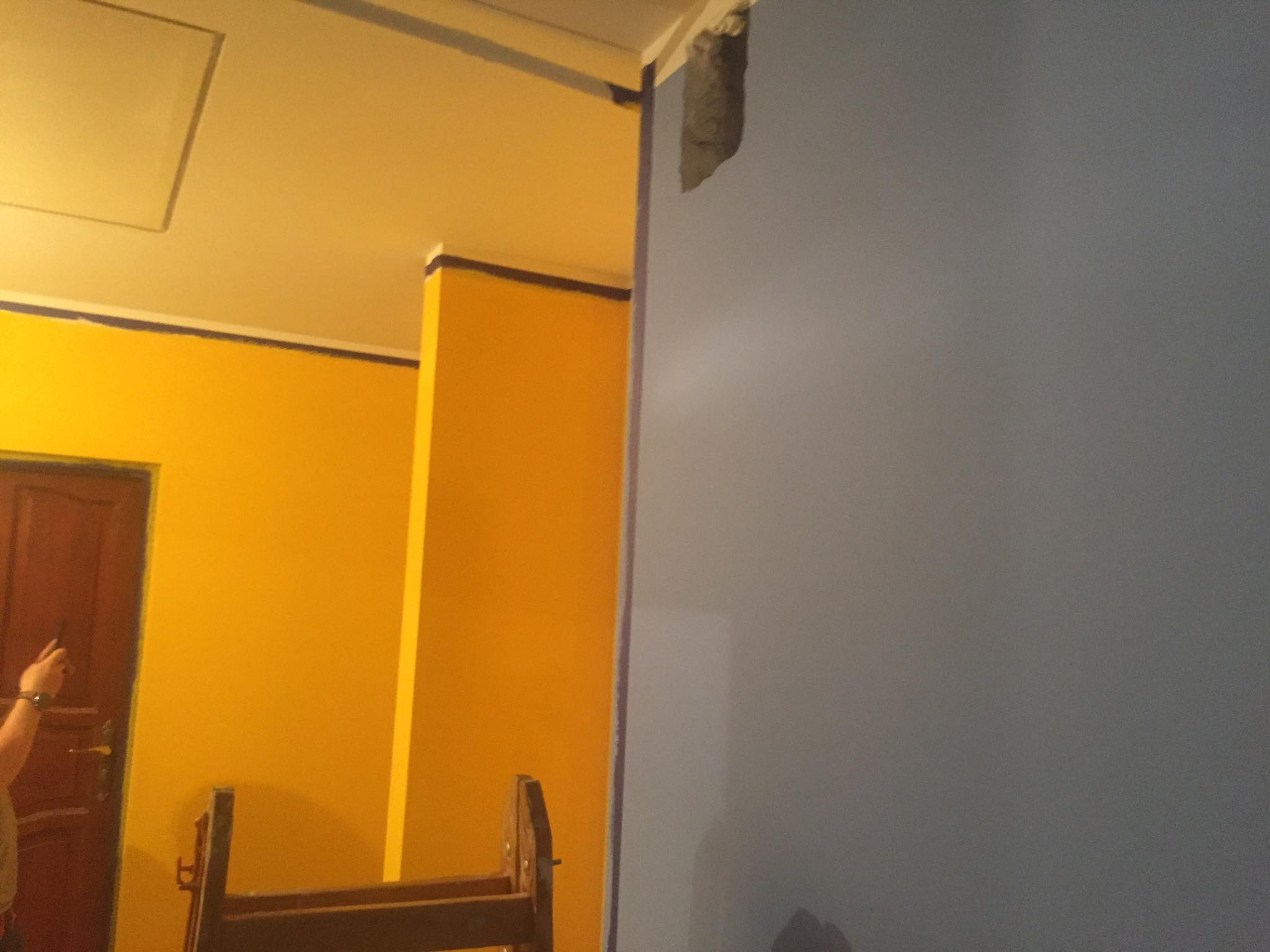 Ściana w trakcie malowania na szaro, z widocznym fragmentem żółtej ściany, zabezpieczonej taśmą malarską, oraz drabina malarska w pomieszczeniu.
