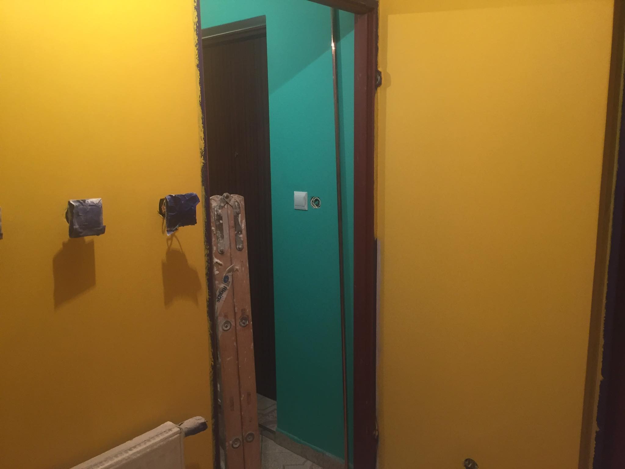 Pokój w trakcie malowania: żółte ściany, zabezpieczone gniazdka, drabina oparta o ścianę z pomalowaną na turkusowo ścianą i futryną drzwi.
