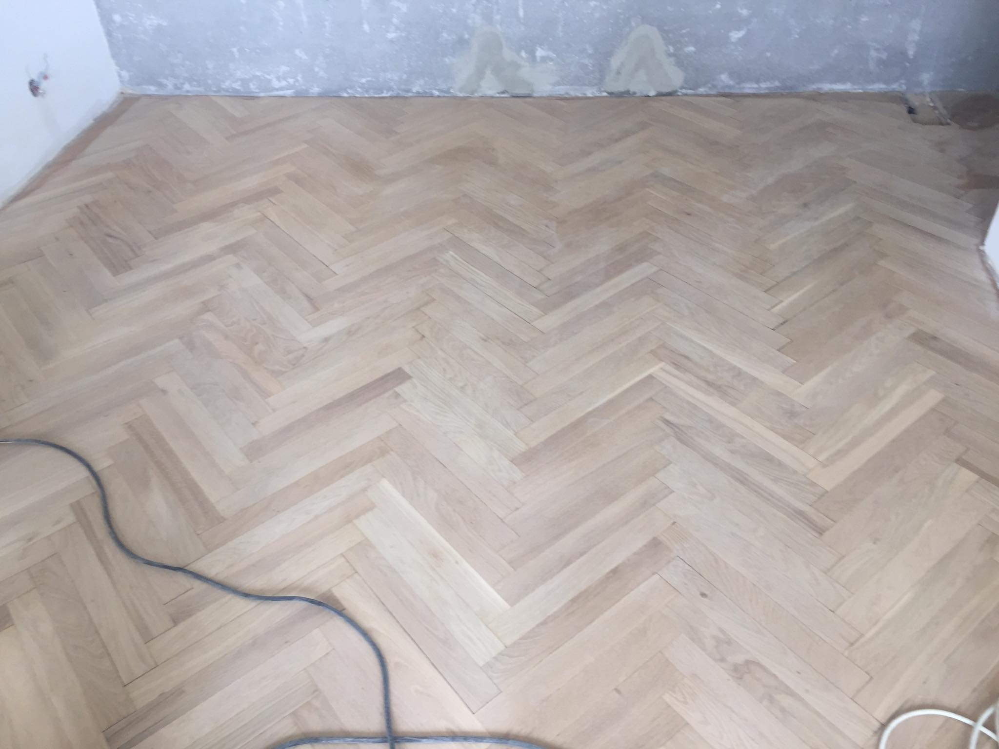 Ułożona parkietowa podłoga w jodełkę, jasne drewno, ściana w tle częściowo wyszpachlowana, widoczny kabel urządzenia do obróbki drewna.