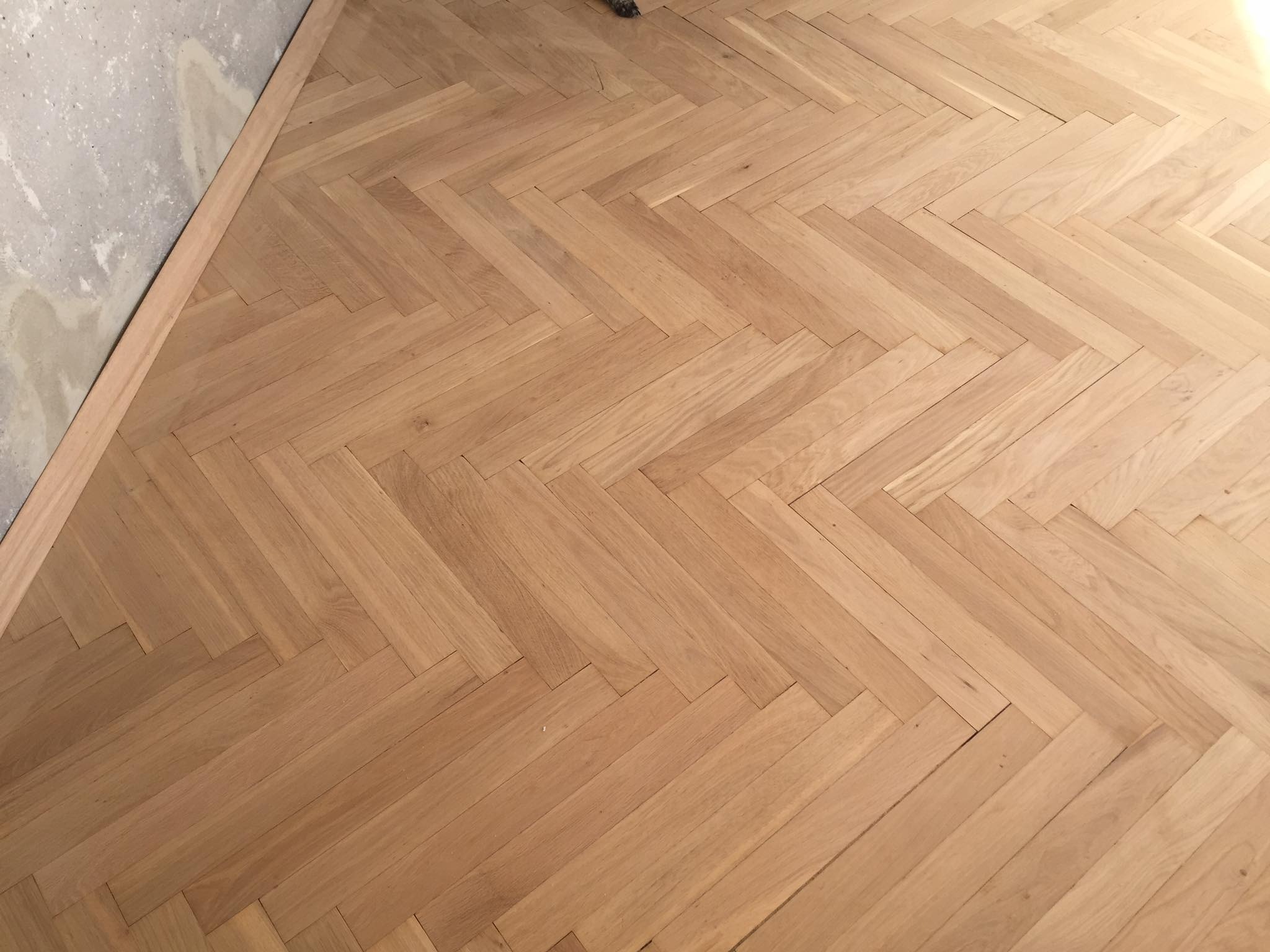 Ukończona podłoga z parkietu ułożonego w jodełkę, jasne drewno, widoczna listwa przypodłogowa i fragment surowej ściany w rogu.