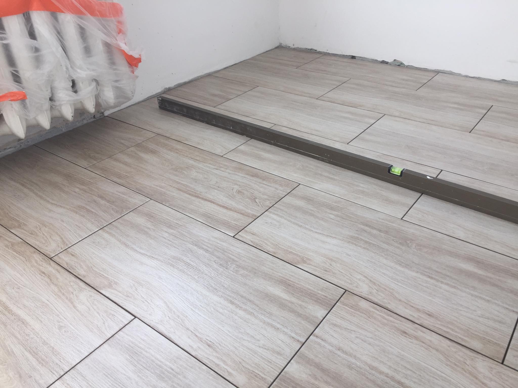 Układanie podłogi z płytek imitujących drewno w rogu pomieszczenia, poziomica spoczywa na płytkach, grzejnik zabezpieczony folią.