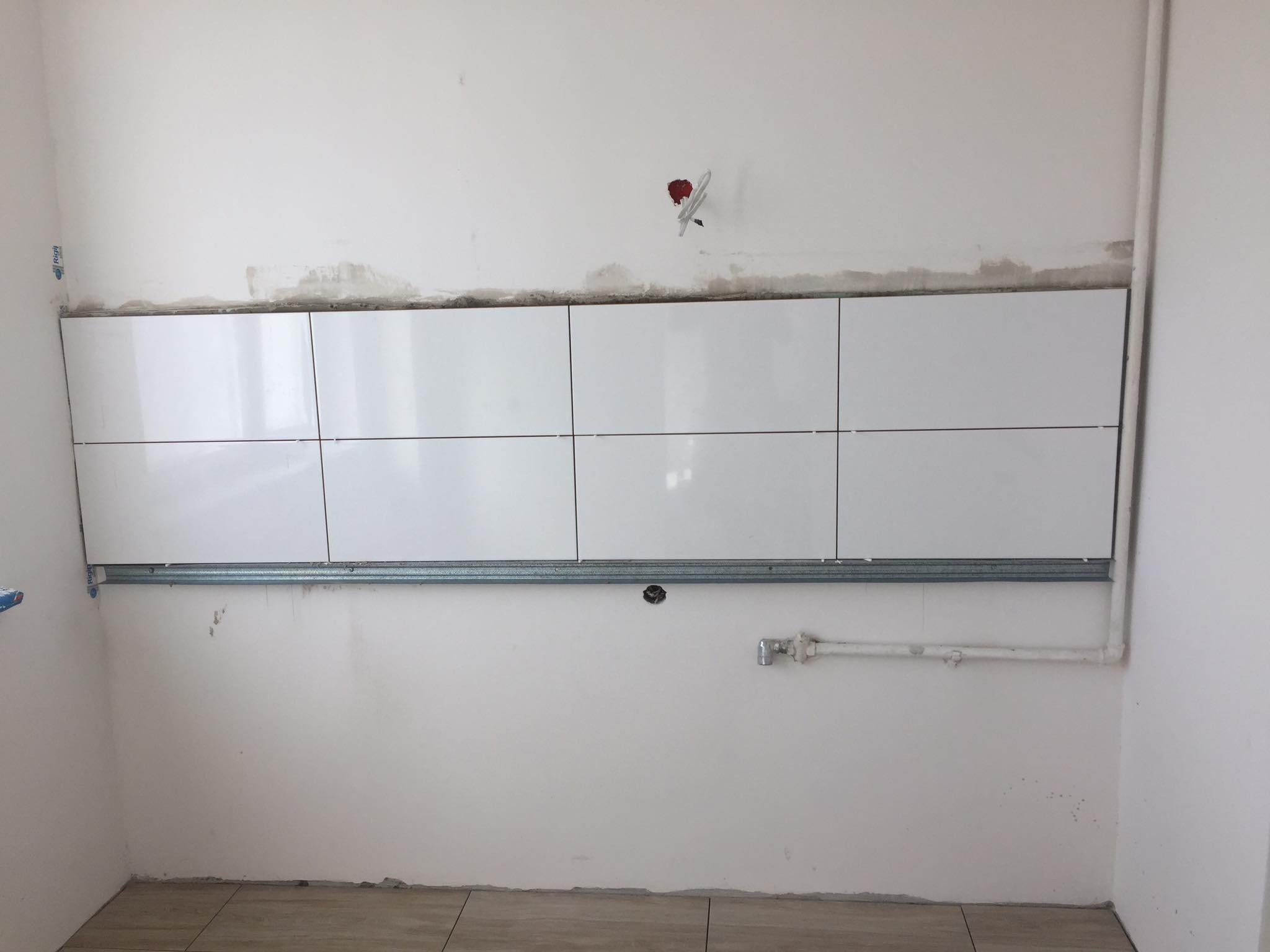 Białe, błyszczące płytki ułożone w rzędzie na ścianie, podparte metalową szyną montażową, widoczne ślady po poprzednich pracach na ścianie, przygotowanie do dalszych prac wykończeniowych.