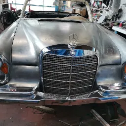 Przód klasycznego Mercedesa w trakcie renowacji, z widocznymi śladami szlifowania i przygotowania do lakierowania, w warsztacie z narzędziami w tle.