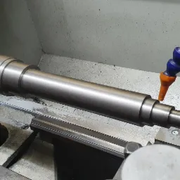 Metalowy wałek obrabiany na tokarce, widoczny system chłodzenia z niebieskim wężykiem i pomarańczową dyszą.