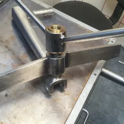 Metalowy element z uchwytem i ostrzem, prawdopodobnie część maszyny, leżący na metalowym stole warsztatowym, obok gaśnica i inne metalowe elementy