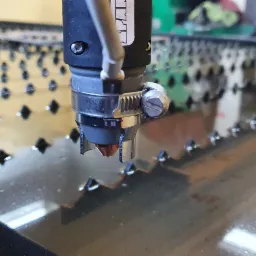 Zbliżenie na głowicę roboczą maszyny CNC podczas cięcia metalu, widoczne iskry i metalowa płyta z wyciętymi otworami w tle.