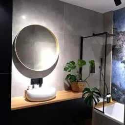 Nowoczesna łazienka z szarymi płytkami, okrągłym lustrem z podświetleniem, białą umywalką nablatową na drewnianym blacie oraz rośliną Monstera w wiklinowym koszu obok wanny z czarną kabiną...