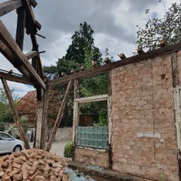 Demontaż ściany z cegły i konstrukcji drewnianej budynku w trakcie remontu, widoczne gruzowisko ceglane i samochód w tle.