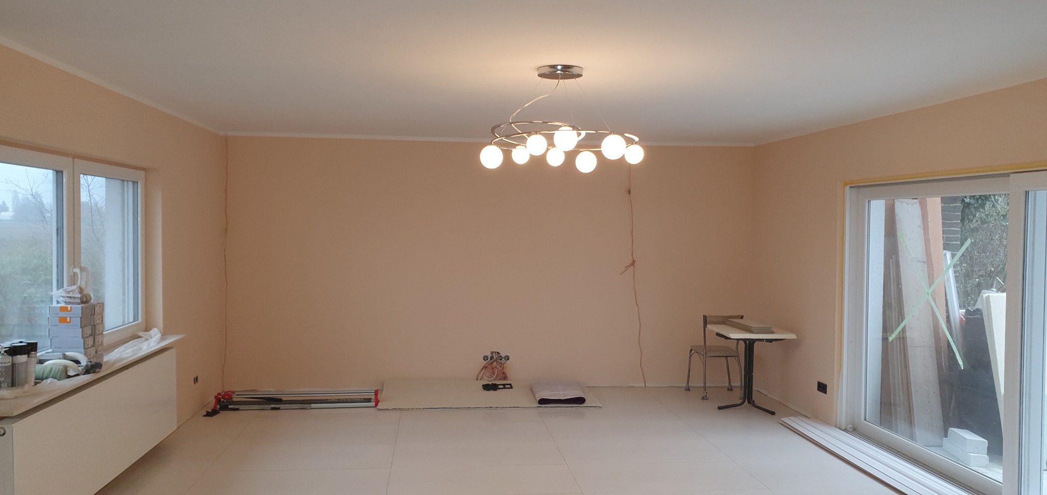 Wnętrze pokoju po malowaniu ścian na brzoskwiniowy kolor, widoczny nowoczesny żyrandol z okrągłymi żarówkami, częściowo ułożona podłoga z jasnych paneli, okno z narzędziami i materiałami malarskimi...