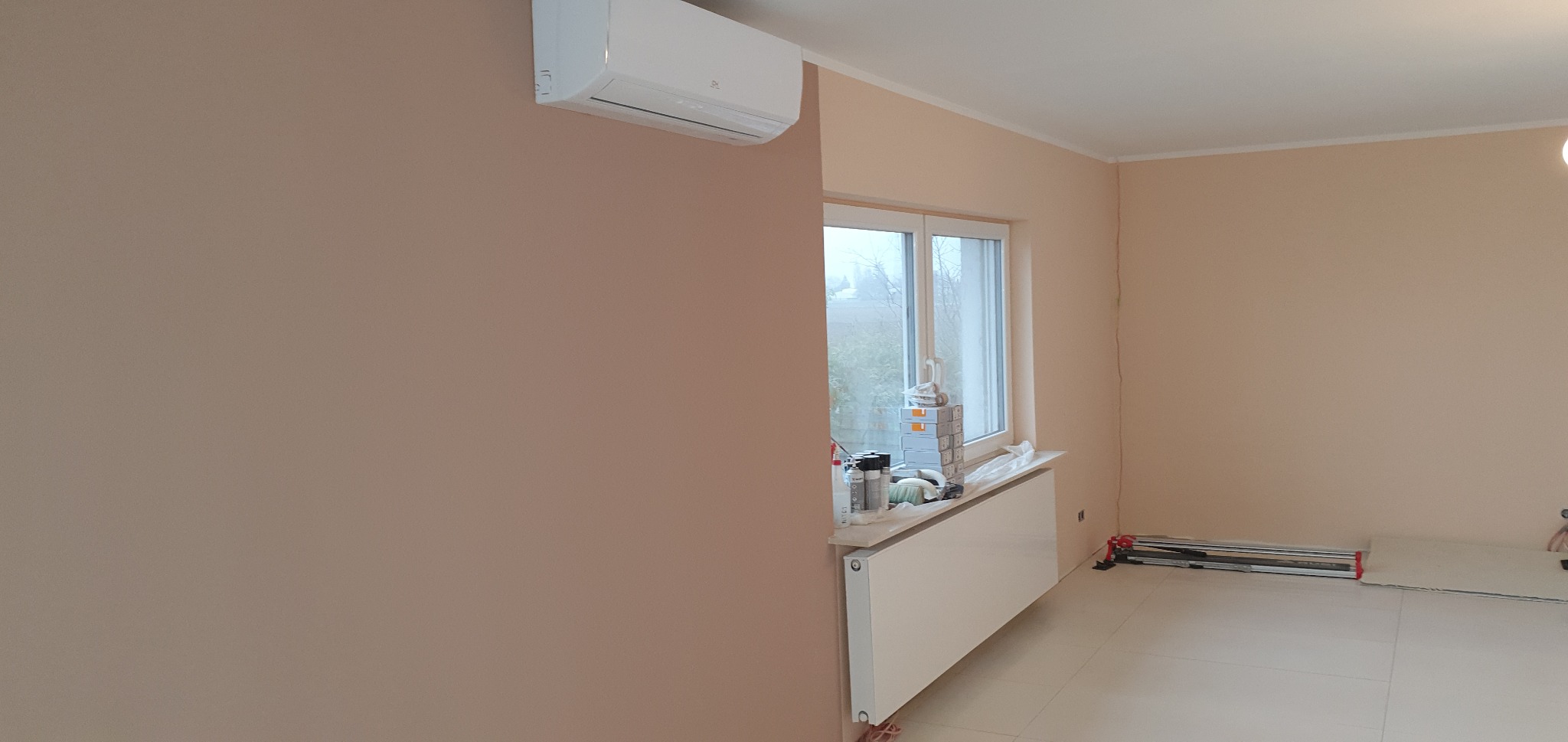 Pomieszczenie po malowaniu na kolor pudrowy róż z zamontowanym klimatyzatorem, widoczne narzędzia i materiały malarskie przy oknie, oraz maszyna do cięcia płytek na podłodze.
