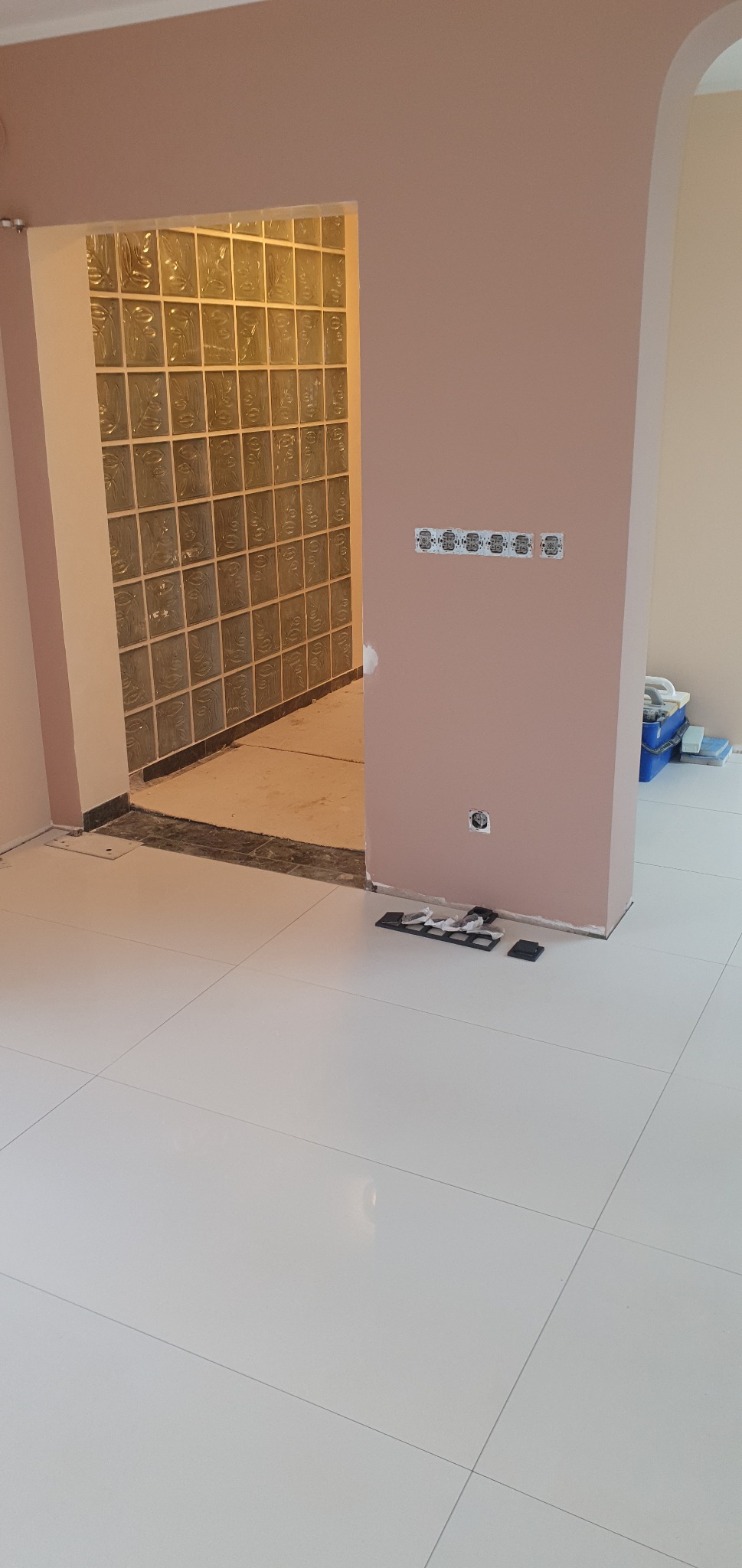 Wnętrze w trakcie prac wykończeniowych: ściana pomalowana na różowo z zamontowanymi puszkami elektrycznymi bez osprzętu, fragment ściany z luksferami, biała podłoga z płytek, narzędzia na podłodze.