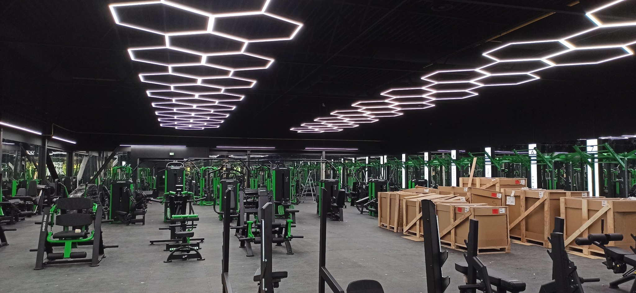 Nowoczesna siłownia z heksagonalnym oświetleniem LED na suficie, zielonymi maszynami do ćwiczeń i kartonami w tle. Widok na przestronne wnętrze.